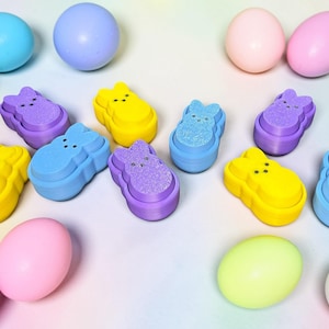 May include: An assortment of colorful Easter decorations, including pastel-colored eggs and bunny-shaped candies. The eggs are in shades of pink, yellow, blue, green, and white. The bunny candies are yellow, purple, and blue with glitter accents.