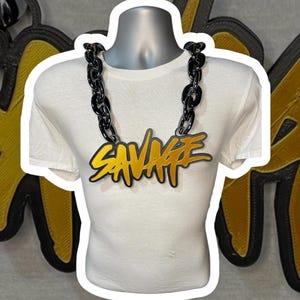 May include: A black and gold chain necklace with the word "SAVAGE" in a yellow and black design. The necklace is displayed on a white t-shirt.