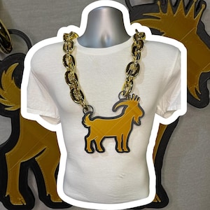 May include: Gold-colored chain necklace with a large pendant featuring a goat silhouette. The pendant is outlined in black and has a gold-colored interior. The necklace is displayed on a white t-shirt.