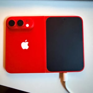 May include: A rectangular red charging device featuring a white Apple logo on the left side, a camera-like module with two lenses and a light, and a large black panel on the right side. A white charging cable is plugged into the bottom edge.