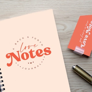 May include: A spiral-bound journal with the words "A Back & Forth Love Notes Journal" in orange and a matching tag that reads "You have a new Love Note." A gold pen rests on a wooden surface.