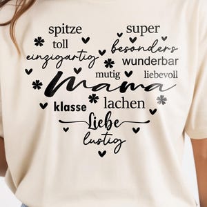 May include: A cream-coloured t-shirt featuring a black heart-shaped design with the German word "Mama" at its centre. The design is surrounded by words such as "spitze", "toll", and "super", with small hearts and flower shapes.