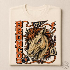 Puede incluir: Una camiseta color crema con un gráfico de un caballo con crin, el texto BRONCOS, LET'S GO! y #1. El diseño incluye un megáfono, una herradura, un rayo y una estrella en tintas de colores naranja, azul y negro.