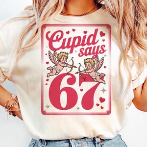 Könnte beinhalten: Cremefarbenes T-Shirt mit Valentinstags-Grafik. Der rosafarbene Aufdruck zeigt "Cupid says 67" zusammen mit Illustrationen von Amorfiguren mit Pfeil und Bogen, Herzen und Sternen. Das Design ist umrandet.