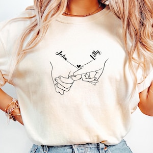 May include: A cream-coloured t-shirt with a black line drawing of two hands holding pinky fingers, with a heart between them. The names "John" and "Lilly" are written above the hands in a script font.