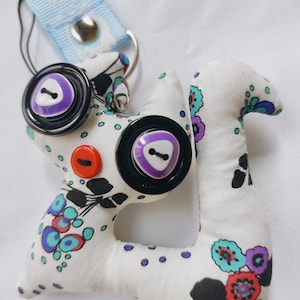 May include: A handmade fabric keychain in the shape of a stylized animal. The white fabric is patterned with colorful floral designs and purple polka dots. Large black buttons with purple centers and an orange button are sewn on the front. A light blue strap is attached.