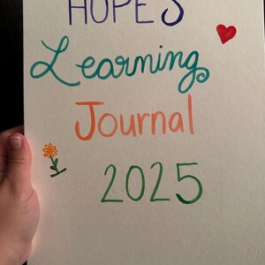 May include: A spiral-bound notebook with the words "HOPE'S Learning Journal 2025" written in colorful script. A red heart and a small flower are drawn on the page. The notebook is held by a hand.