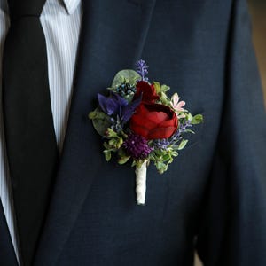 May include: A navy blue suit with a black tie and a floral buttonhole. The buttonhole includes a large red flower, purple accents, and greenery. The suit is tailored, and the shirt is white with thin vertical stripes.