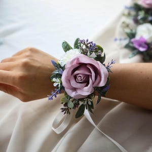 Op de afbeelding: Een polscorsage met een grote mauve roos, witte accenten en takjes paars en groen blad. Een wit lint bevestigt het bloemstuk. Extra corsages zijn zichtbaar op de achtergrond.