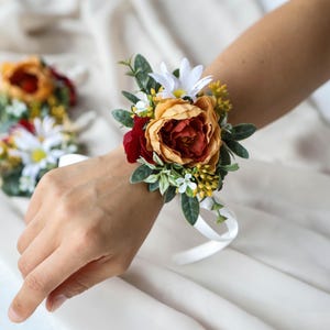 May include: A wrist corsage with a prominent orange and red flower, complemented by white daisies, green foliage, and yellow details. A white ribbon secures the floral arrangement. Other corsages are visible in the background. The arrangement is suitable for weddings.