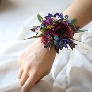 Op de afbeelding: Een polscorsage met een dieprode roos omringd door paarse, blauwe en groene bloemen en bladeren. Het bloemstuk is vastgemaakt met een wit lint. Perfect voor bruiloften, gala's of speciale evenementen.