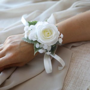 Peut inclure: Un bracelet de corsage floral blanc avec une grande rose entourée de petites fleurs blanches et de feuilles vertes. Le corsage est attaché avec un ruban blanc. Parfait pour les mariages et les événements.