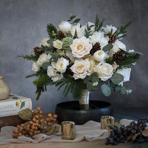 Puede incluir: Un exuberante ramo de rosas blancas, piñas y vegetación. El arreglo está en un jarrón negro, con candelabros dorados y pequeños candeleros dorados. También se ve un libro titulado "Wild Flowers".