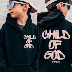 Pode incluir: Moletom preto com a frase "CHILD OF GOD" em rosa claro na frente e atrás. A parte de trás do moletom também inclui "PSALM 23". O moletom tem um bolso frontal e um capuz.