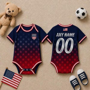 May include: A navy and red baby bodysuit with a star pattern and the text "USA" on the front. The back of the bodysuit has the text "ANY NAME" and the number "00". The image also includes a teddy bear, a football, and baby shoes.