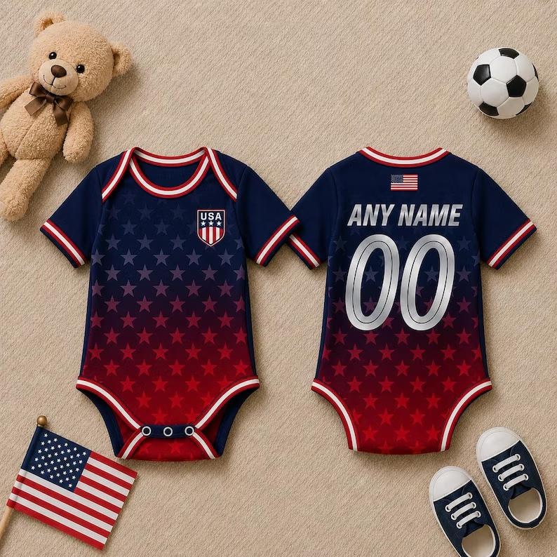 May include: A navy and red baby bodysuit with a star pattern and the text "USA" on the front. The back of the bodysuit has the text "ANY NAME" and the number "00". The image also includes a teddy bear, a football, and baby shoes.