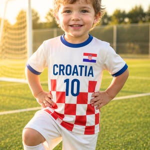 May include: A child's white football kit featuring the word "CROATIA" and the number "10". The kit has a red and white chequered design and a Croatian flag. The child is standing on a green football pitch.
