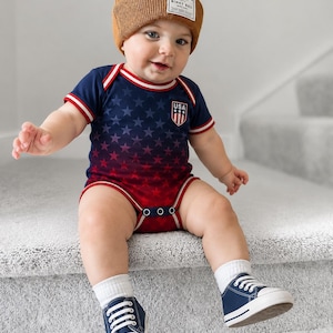 May include: A baby in a navy and red USA-themed onesie with a star pattern, a brown beanie, white socks, and navy sneakers with white laces. The onesie features a USA crest. The beanie has a label.