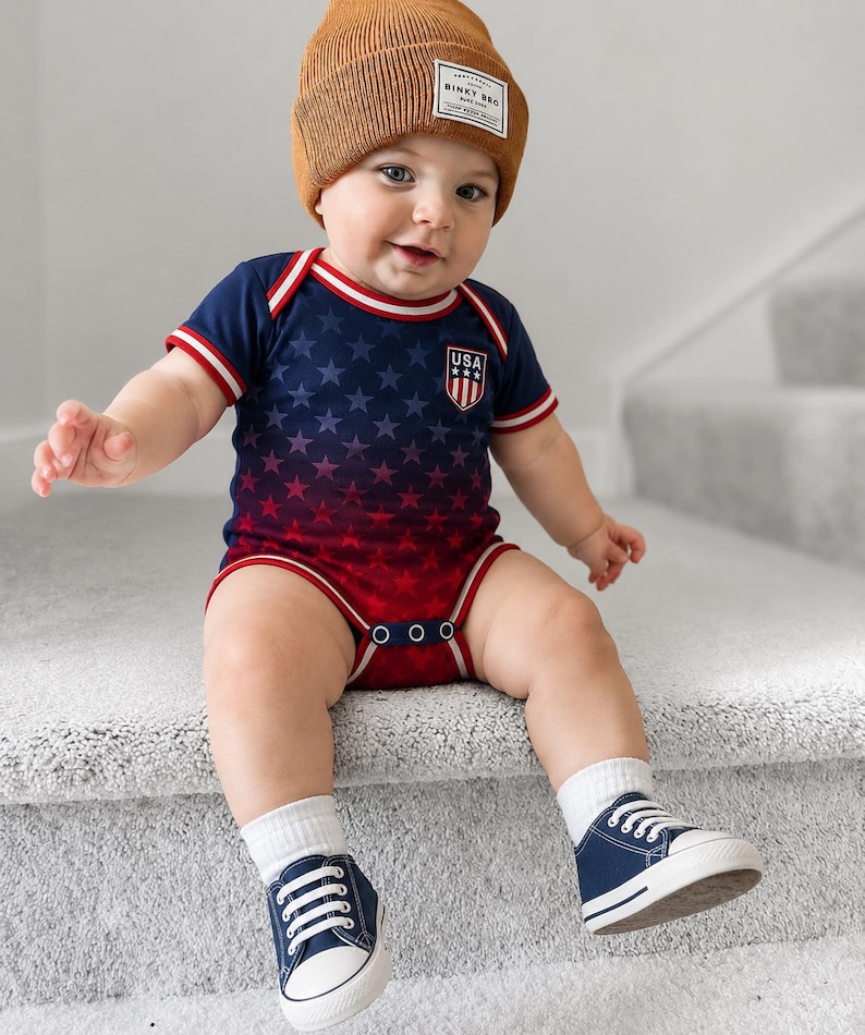 May include: A baby in a navy and red USA-themed onesie with a star pattern, a brown beanie, white socks, and navy sneakers with white laces. The onesie features a USA crest. The beanie has a label.