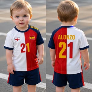 May include: A toddler wearing a white, red, and navy blue soccer jersey and shorts. The jersey has the number 21 in yellow and red, and the name "ALONZO" on the back. The jersey also features the flags of England and Spain.