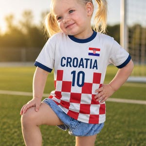 Puede incluir: Una camiseta blanca con ribetes azul marino que muestra la palabra "CROATIA" y el número "10" en azul marino. La camiseta tiene un patrón de cuadros rojos y blancos y una bandera croata. El niño lleva pantalones cortos vaqueros.
