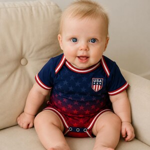 May include: A baby in a navy and red star-patterned bodysuit with a "USA" emblem. The bodysuit has white and red trim. The baby is sitting on a beige cushion. The outfit is buttoned at the bottom.