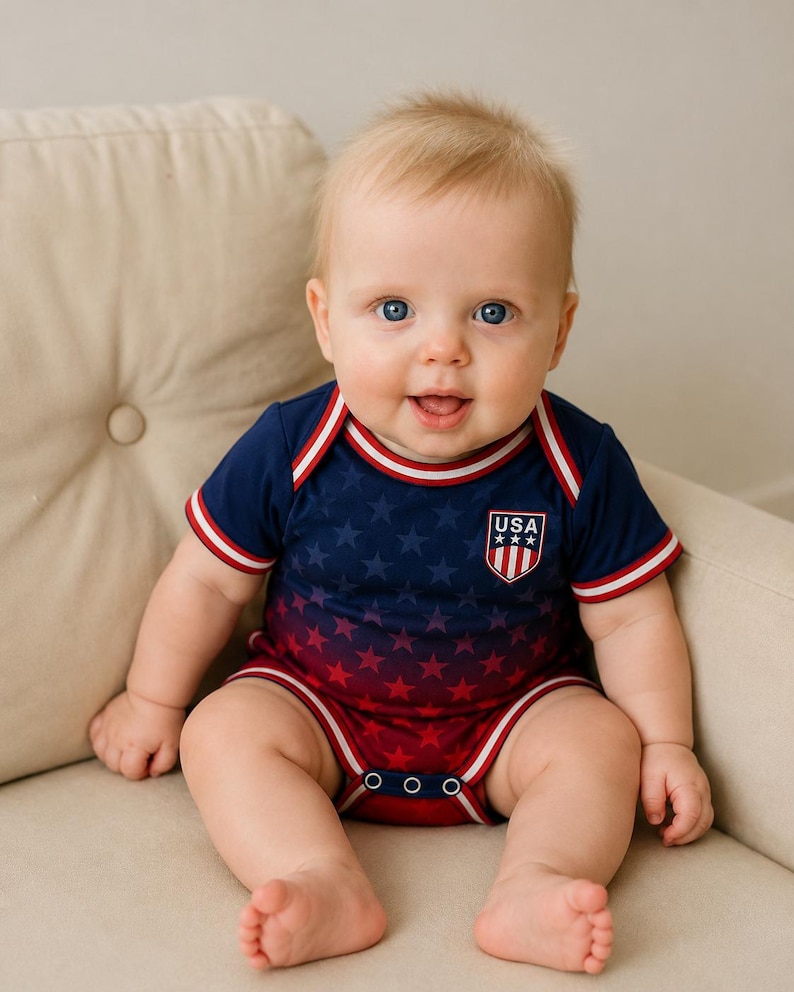 May include: A baby in a navy and red star-patterned bodysuit with a "USA" emblem. The bodysuit has white and red trim. The baby is sitting on a beige cushion. The outfit is buttoned at the bottom.
