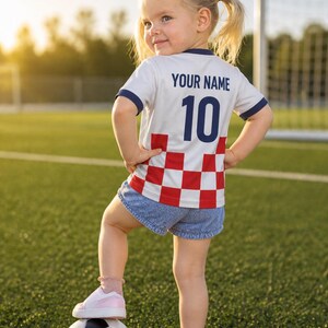 May include: A child's football shirt, white with blue trim, the number 10, and the text "YOUR NAME" on the back. The shirt has a red and white chequered pattern at the bottom. The child is standing on a football.