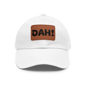 May include: A white baseball cap featuring a rectangular brown patch centered on the front. The patch displays the word DAH! in bold, black, rounded lettering. The cap has a curved brim and a structured crown with stitched eyelet details.