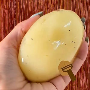 Puede incluir: Una patata grande, lisa y amarilla sostenida en una mano. La patata tiene pequeñas manchas oscuras y una superficie brillante. Una ilustración de dibujos animados de un pelador de patatas está al lado de la patata.