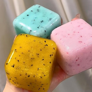 以下が含まれることがあります： 黄色、ピンク、ティール色の3色の正方形の石鹸。石鹸は半透明で、濃い斑点があります。石鹸は手で持たれています。