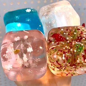 Può includere: Quattro oggetti decorativi trasparenti di forma quadrata. Uno è blu con accenti di fiocchi di neve bianchi, un altro è rosa con accenti di fiocchi di neve bianchi, un terzo è bianco con accenti argentati e l'ultimo è pieno di decorazioni rosse, verdi e dorate.