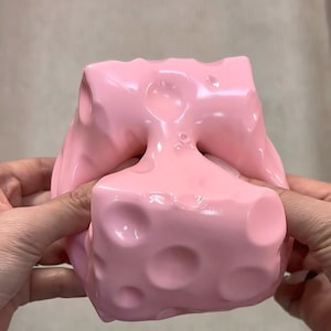 Könnte beinhalten: Ein weiches, biegsames, hellrosa Quetschspielzeug mit einem Design, das an Schweizer Käse erinnert. Das Objekt hat eine glatte, glänzende Oberfläche mit verschiedenen kreisförmigen Vertiefungen unterschiedlicher Größe auf seiner flexiblen Form.