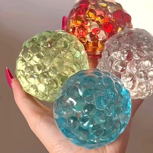 May include: A hand displays four colourful water beads. The beads are in shades of green, red, clear, and blue. Each bead is composed of many small, clear spheres. The beads are a vibrant and eye-catching sensory toy, ideal for children.