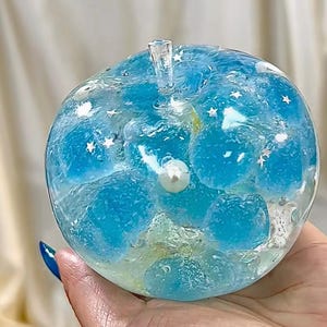 May include: A clear, apple-shaped decorative object filled with blue spheres and white star accents. A pearl is embedded in the center. The apple has a clear stem. The overall design is whimsical and artistic.