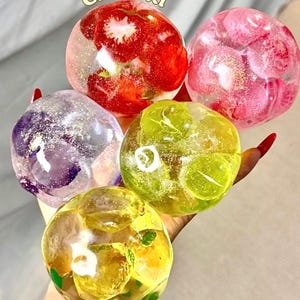 Può includere: Cinque fermacarte sferici in resina trasparente contenenti fettine di frutta e glitter. Le sfere presentano fragole rosse, lamponi rosa, uva viola, fettine di lime verde e fettine di limone giallo, sospese in un materiale lucido e trasparente.