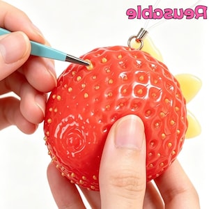 Peut inclure: Un objet rouge vif en forme de fraise avec des graines jaunes et une boucle de suspension transparente. Une pince bleu clair est utilisée pour placer une petite graine jaune dans un trou à la surface. Le mot Reusable est écrit en rose en haut.