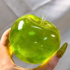 Peut inclure: Un objet décoratif en forme de pomme vert translucide avec une tige transparente. L'objet est tenu dans une main, et la pomme a une finition brillante. Le thème général est celui des fruits décoratifs.