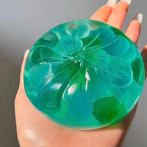 May include: A round, translucent soap bar with a floral design. The soap is a mix of teal and green, with lighter blue accents. The soap is held in a hand, showcasing its shape and colour variations. The soap appears to be handmade.