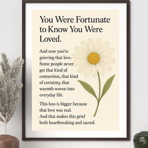 Puede incluir: Impresión artística enmarcada con el texto "You Were Fortunate to Know You Were Loved". Se presenta una ilustración de una margarita. La impresión está en un marco negro y descansa sobre una estantería de madera.