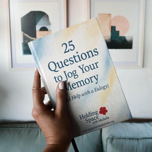 Peut inclure: Un livre intitulé "25 Questions to Jog Your Memory" est tenu devant un mur avec des œuvres encadrées. La couverture du livre présente un design aquarelle avec des teintes bleues et jaunes. Le texte comprend "and Help with a Eulogy" et "Holding Space with Michelle."