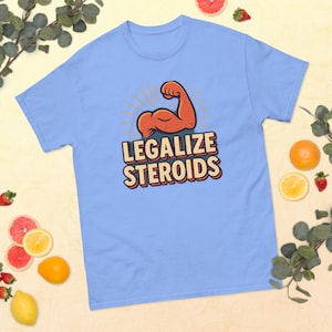 Puede incluir: Camiseta azul claro con el texto "LEGALIZE STEROIDS" en una fuente retro. El diseño incluye un músculo de brazo de dibujos animados naranja y marrón flexionándose, con rayos de sol detrás.