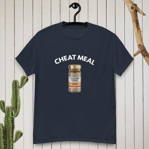 Pode incluir: Camiseta azul marinho com a frase "CHEAT MEAL" em branco acima de um gráfico de um frasco de suplemento. O frasco tem um rótulo castanho e branco com o texto "TESTOSTERONE CYPIONATE".