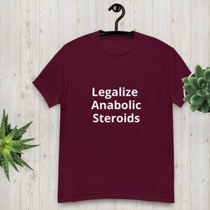 Pode incluir: Uma t-shirt bordô com o texto branco "Legalize Anabolic Steroids". A t-shirt está pendurada num cabide preto sobre um fundo claro com efeito de madeira. Uma pequena planta suculenta é visível à direita.