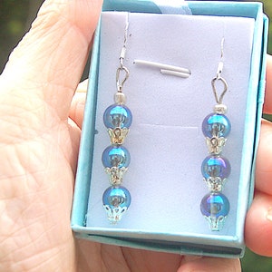Op de afbeelding: Een paar oorhangers met drie iriserende blauwe kralen aan elke oorhanger. De oorhangers zitten in een klein blauw doosje.