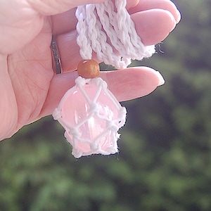 May include: A pink crystal pendant encased in a white macrame net, suspended from a white cord. A small wooden bead sits above the crystal. The background is a blurred green.