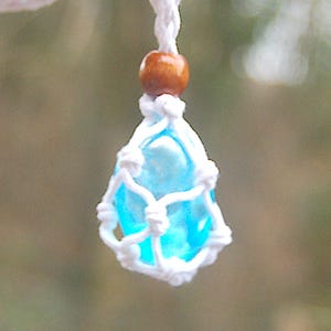 Puede incluir: Un cristal azul claro está envuelto en un nudo de macramé blanco. El cristal está suspendido de un cordón blanco con una cuenta de madera marrón en la parte superior.