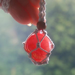 Può includere: Un ciondolo artigianale in pietra preziosa rossa, racchiuso in una rete di macramè bianca, sospeso a un cordino bianco. Una perlina di legno accentua il cordino. Il ciondolo è tenuto da una mano.