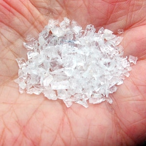 Puede incluir: Un puñado de piezas de plástico transparentes, de forma irregular y translúcidas. Los fragmentos son pequeños y parecen estar dispersos sobre la palma de una mano. Las piezas tienen un aspecto cristalino.