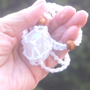 May include: A white macrame crystal holder with a clear, iridescent crystal sphere. The holder has wooden beads and is held in a hand. The background is blurred green.